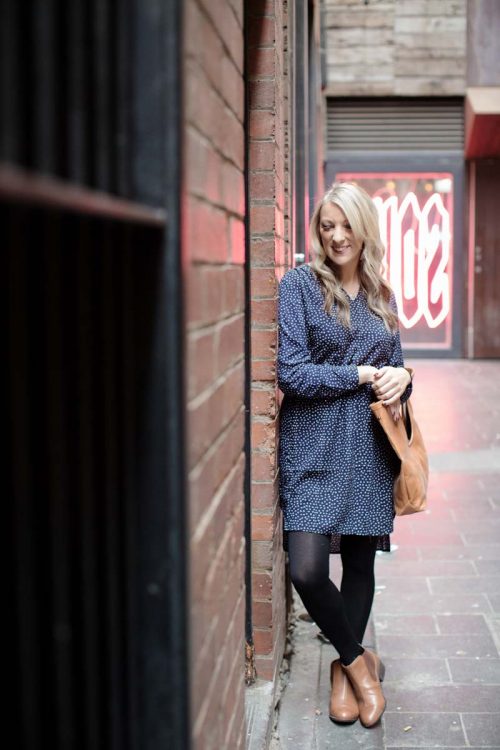 Blonde woman leaning against a wall, smiling with her bag