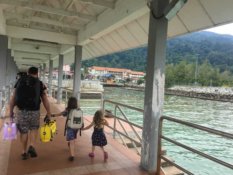 A father holding hands with his daughter who's holding her little sister's hand on a dock overlooking the beautiful blue water as a boat passes